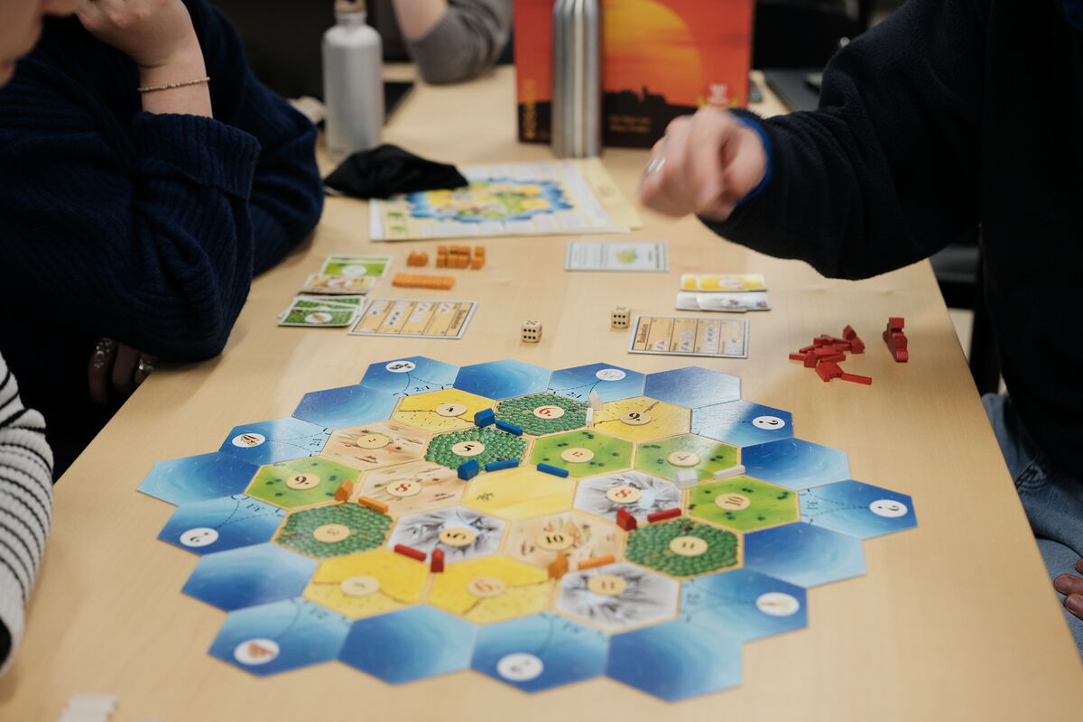 People sitting around tables in a T-shape, playing the Catan boardgame, three laptops are on the table as well.